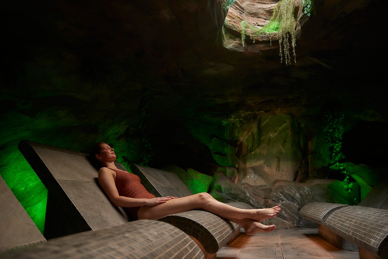 A woman reclines on a tiled lounge chair, eyes closed. She relaxes in a cave-like spa room with green lighting and a circular skylight above, plants dangling from the opening.